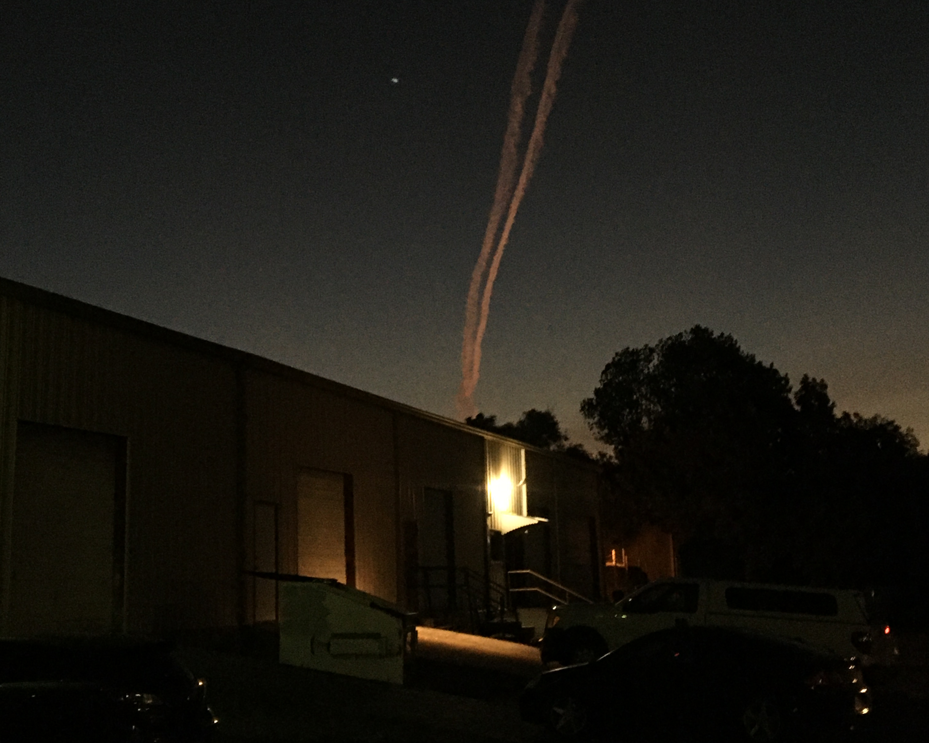 a jet's contrail is faintly illuminated by the not yet risen sun above dark shadows of industrial warehouses and trees