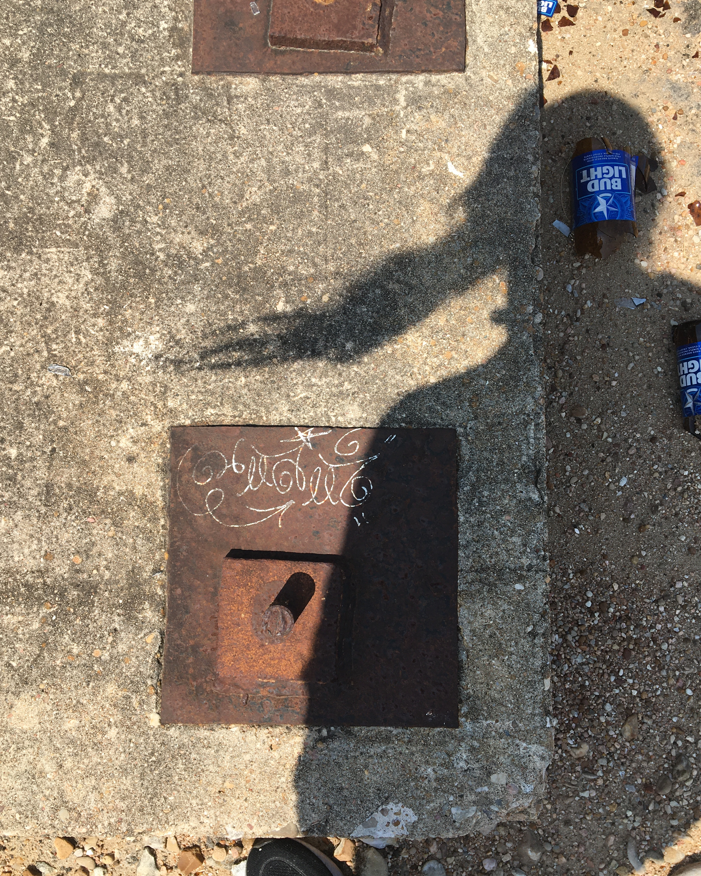 looking down at a cement block with rusted fixtures, a shadow of the photo taker's long hair blowing in the wind cast over it, and several shattered bud lite cans strewn around