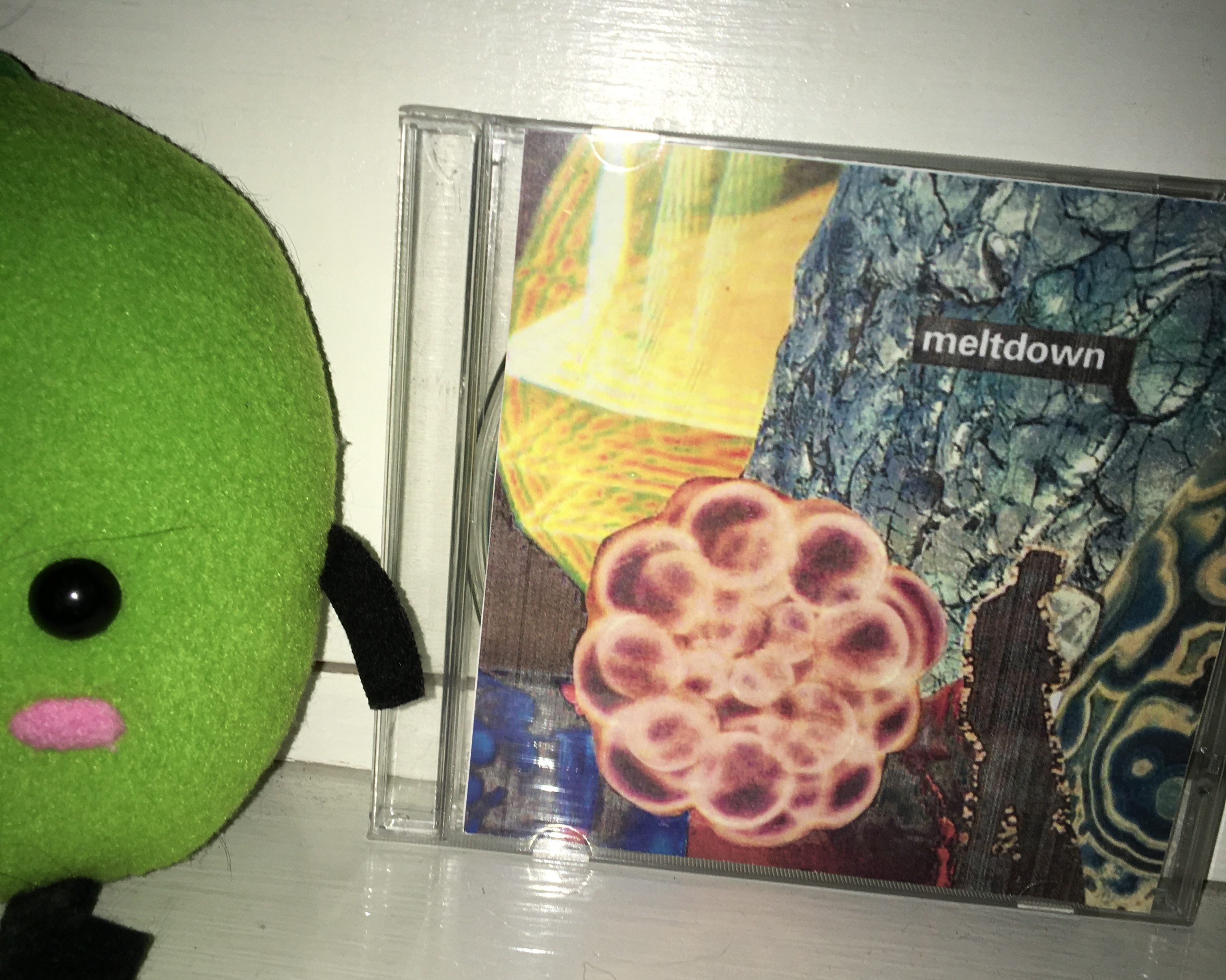 a close up, flash-photo of a cd case with custon album cover set on the shelf of a book case next to a green junimo plush barely out of frame