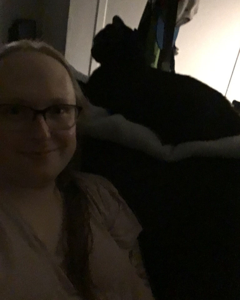 blonde girl sitting on sofa in dark room with a black cat perched above on the top of the couch