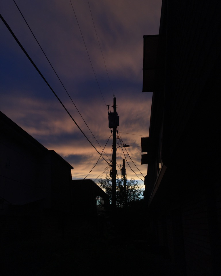 foregroud of buildings & power lines in shadow, amongst a background of soft clouds illuminated yellow & blue just after a storm