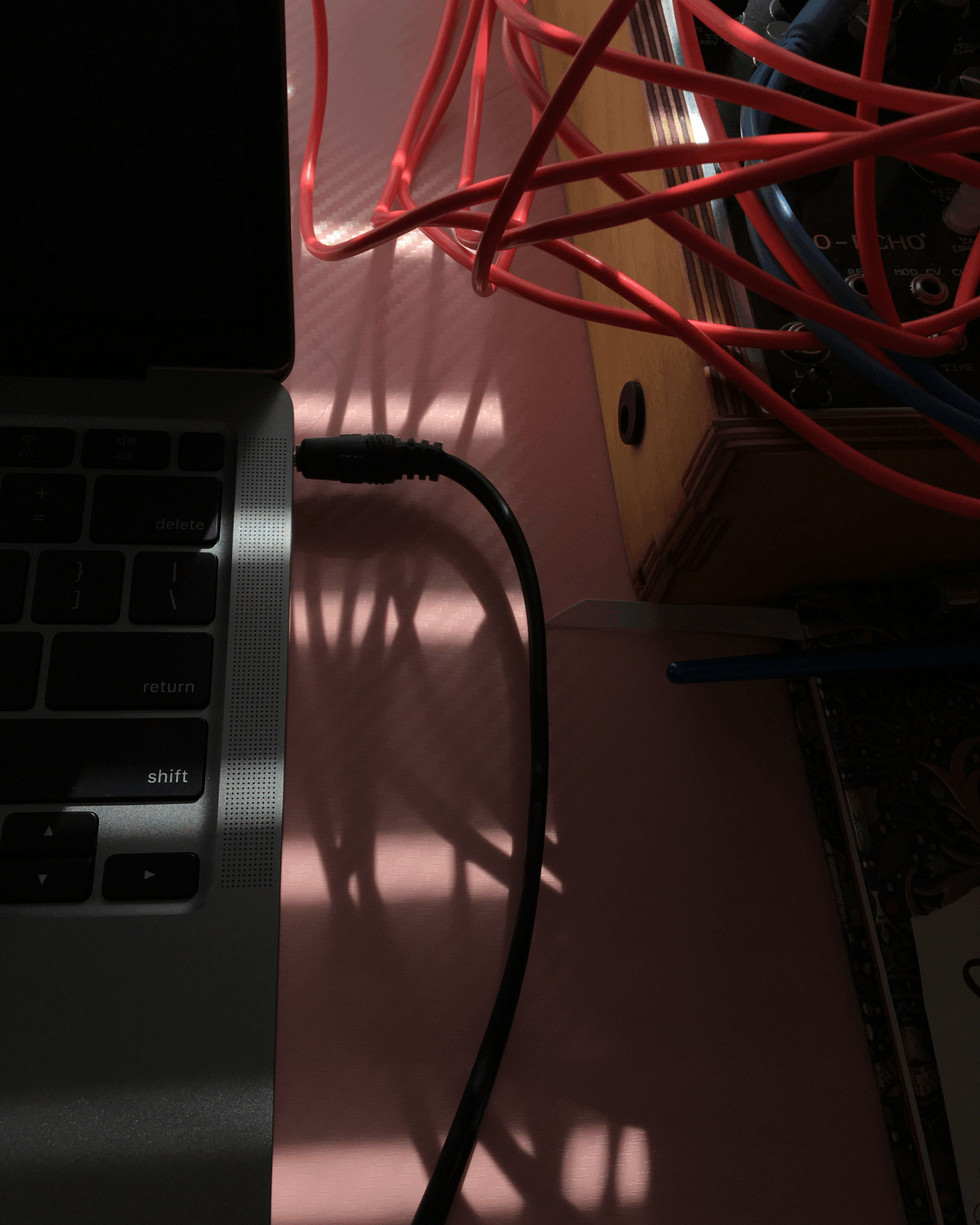 thin rays of sun sun shining through horizontal blinds, casting shadows from a tangle of synthesizer patch cables on to a macbook next to them on a pink desk,